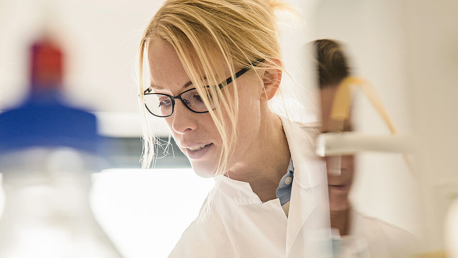 A female scientist with blonde hair and glasses, wearing a white lab coat, working in a laboratory setting.