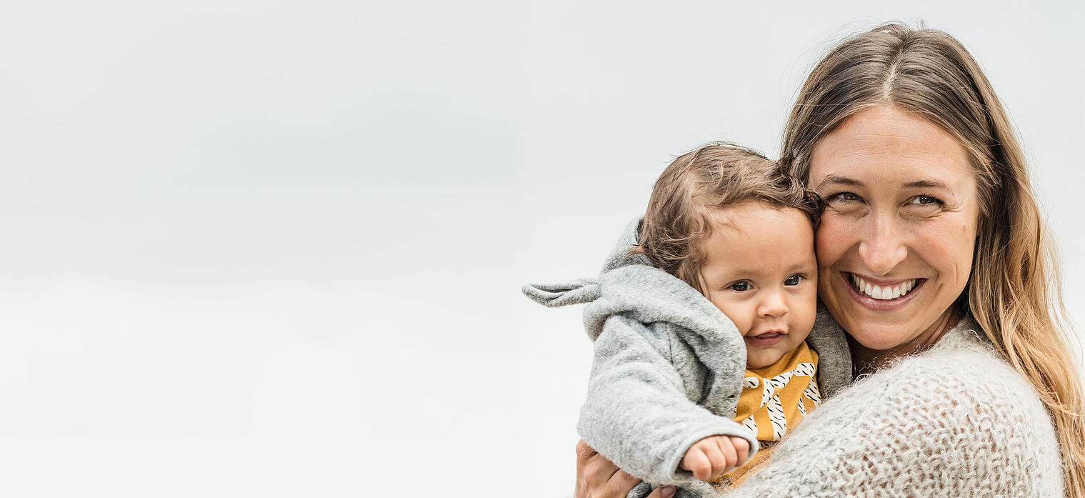 Smiling woman holding a happy baby, both dressed warmly in cozy clothing, against a neutral background.