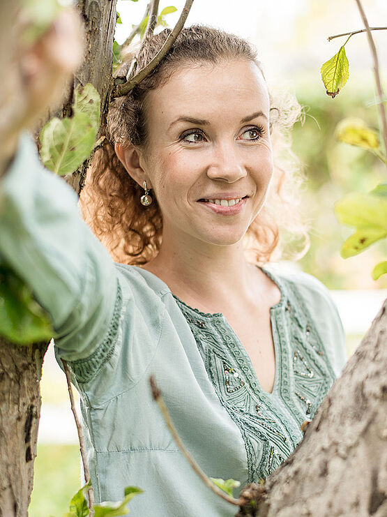 Smiling woman with curly hair wearing a light green blouse, standing amidst tree branches and greenery.